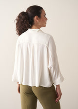 Woman wearing a creamy white blouse and green tapered trousers against a plain background