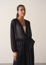 Women wearing a long sleeve sheer sparkle draped top with cream satin camisole and black satin trousers, on a beige background