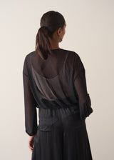 Women wearing a long sleeve sheer sparkle draped top with satin camisole and black satin trousers, facing towards a beige wall