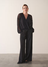 Woman wearing a black sheer top and black wide leg trousers against a plain background