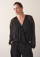 Woman wearing a black sheer top and black wide leg trousers against a plain background