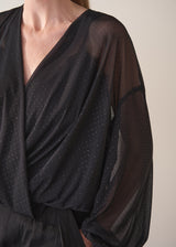 Woman wearing a black sheer top and black wide leg trousers against a plain background