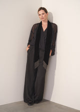 Woman wearing a black sheer top and black wide leg trousers against a plain background