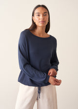 A woman wearing a navy blue long-sleeve top with a wide boat neckline and front drawstring hem, paired with light beige trousers.