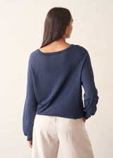 A woman wearing a navy blue long-sleeve top with a wide boat neckline and front drawstring hem, paired with light beige trousers.