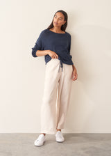 A woman wearing a navy blue long-sleeve top with a wide boat neckline and front drawstring hem, paired with light beige trousers.
