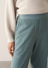 Woman wearing a cream rollneck sweater and aqua cord trousers against a plain background