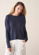 A woman stands against a plain off-white wall wearing a navy blue knit top with a brown tie detail, white wide-leg trousers, and beige slip-on shoes.