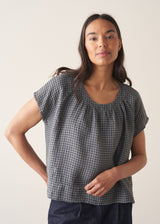 Woman wearing a blue and green check linen top and denim cullottes against a plain background