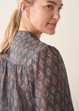 A woman wearing a dark blue long-sleeve blouse with a brown and rust-colored paisley print pattern and light trousers.