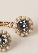 Close up of a pair of vintage style gold drop earrings with pearls surrounding a smokey grey crystal, on a beige background