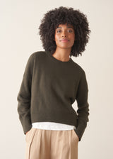 A woman wearing a khaki green crewneck sweater, layered over a white top and beige trousers.