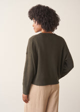 A woman wearing a khaki green crewneck sweater, layered over a white top and beige trousers.