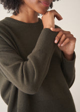 A woman wearing a khaki green crewneck sweater, layered over a white top and beige trousers.