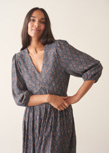 A woman wearing a dark blue midi dress with a brown and beige paisley pattern, featuring puff sleeves and a deep V-neckline, set against a plain beige background.