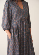 A woman wearing a dark blue midi dress with a brown and beige paisley pattern, featuring puff sleeves and a deep V-neckline, set against a plain beige background.