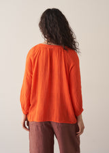 Woman wearing a bright orange cotton blouse and linen trousers against a plain background