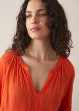 Woman wearing a bright orange cotton blouse and linen trousers against a plain background