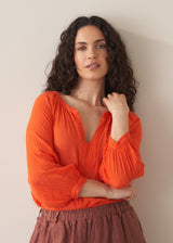 Woman wearing a bright orange cotton blouse and linen trousers against a plain background