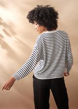 Woman wearing a light knit striped crew neck sweater with black trousers, facing a neutral backgroud