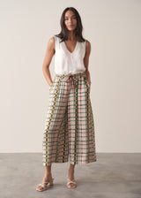 A woman models silk blend wide-leg trousers in a check print, paired with a sleeveless satin top and strappy sandals. She stands in front of a beige background