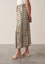 A woman wears silk-blend wide-leg trousers in a check print with a sleeveless satin top and strappy sandals, against a beige background