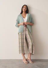 A woman stands against a beige background, wearing a chunky knit cardigan over a white top, paired with silk-blend wide-leg trousers and strappy sandals