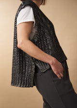 Woman wears a dark grey silver metallic cable knit vest, white short sleeve top and black trousers.