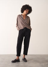 Woman wearing a light brown long-sleeve polo neck sweater and black drawstring trousers against a plain background