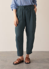 Woman wearing a pair of dark green, tapered leg, ankle-length linen trousers with a blue blouse, against a beige background