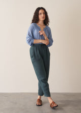 A woman wearing dark green tapered leg, ankle length linen trousers with a blue blouse, against a beige background