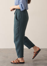 Woman strolling in a pair of dark green, tapered leg, ankle-length linen trousers styled with a blue blouse