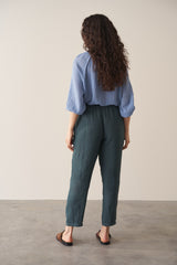 Woman facing a wall, wearing dark green tapered leg, ankle-length linen trousers with a blue blouse, against a beige background