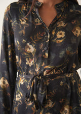 A woman wearing a long-sleeve, dark floral midi dress with a tie waist, set against a plain off-white studio background.
