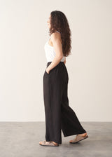 Woman wearing a white tank top and black wide-leg linen trousers against a plain background