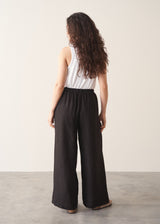 Woman wearing a white tank top and black wide-leg linen trousers against a plain background