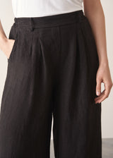 Woman wearing a white tank top and black wide-leg linen trousers against a plain background