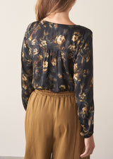 A woman wearing a long-sleeve dark floral blouse with a V-neckline, paired with mustard yellow drawstring barrel leg trousers