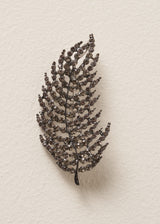 Leaf brooch embellished with smoky-grey crystals, against a beige background