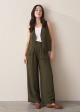 Woman wearing dark green linen blend wide-leg trousers and coordinating waistcoat, styled with brown leather loafers, against a beige background