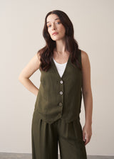 Woman wearing green waistcoat, green high waisted trousers and white vest top against a beige background