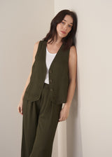 Woman wearing a green waistcoat, green high-waisted trousers and white vest top against a beige background