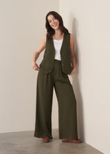 Woman wearing a green waistcoat, green high-waisted wide leg trousers and a white vest top with brown leather loafers against a beige background