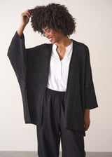 Woman wearing a dark gray cardigan over a white shirt and black trousers against a plain background