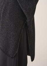 Woman wearing a dark gray cardigan over a white shirt and black trousers against a plain background