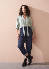 Woman wearing a light green blouse and dark blue jeans against a beige wall.