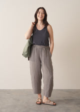 Woman wearing a dark grey tank top and checked linen trousers with sandals, against a plain background