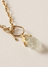 Gold chain necklace with a clear chunky crystal pendant on a beige background