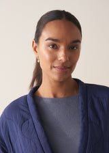 Woman wearing crystal gold heart hoop earrings, with a blue jacket, on a beige background