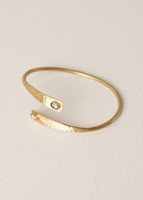 A hammered gold open cuff bangle bracelet with a small clear gemstone accent on each end, displayed on a textured off-white background.